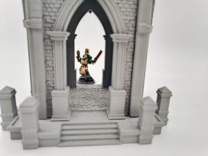 Grey 3D printed Gothic archway tabletop gaming terrain with a painted miniature figure standing in the doorway
