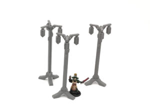 Three grey 3D-printed miniature lampposts on a white background, shown next to a painted figure for scale