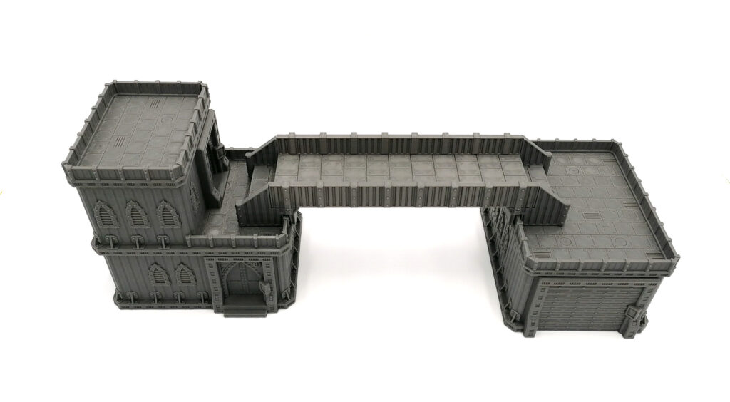 Grey 3D-printed tabletop gaming terrain with two industrial buildings connected by an elevated walkway