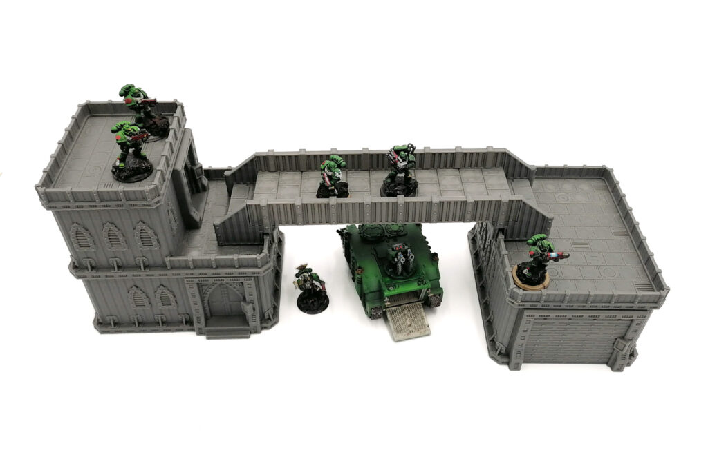 Grey modular sci-fi tabletop terrain with a bridge and buildings, shown with green miniature soldiers and a tank