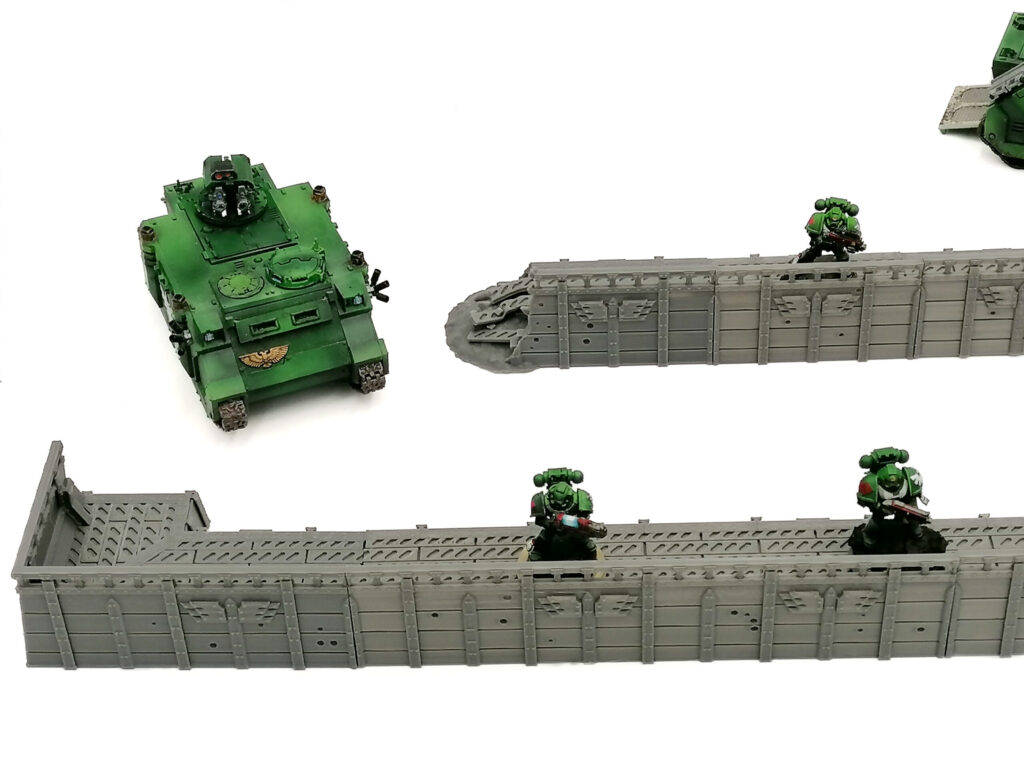 Modular grey 3D-printed defensive walls for tabletop wargaming, shown with miniature green soldiers and a tank