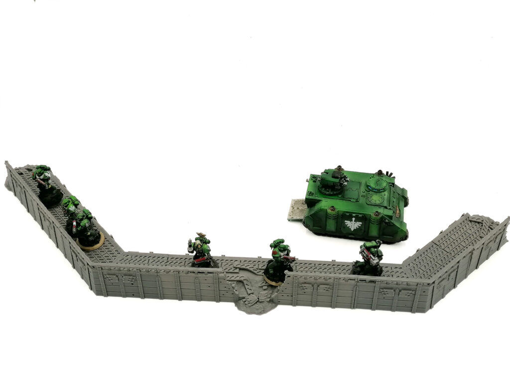 Grey 3D-printed modular defensive wall terrain for tabletop wargaming, shown with miniature figures for scale