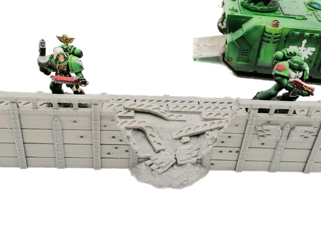 Grey 3D-printed miniature defensive wall with a blasted central breach, shown with sci-fi models for scale