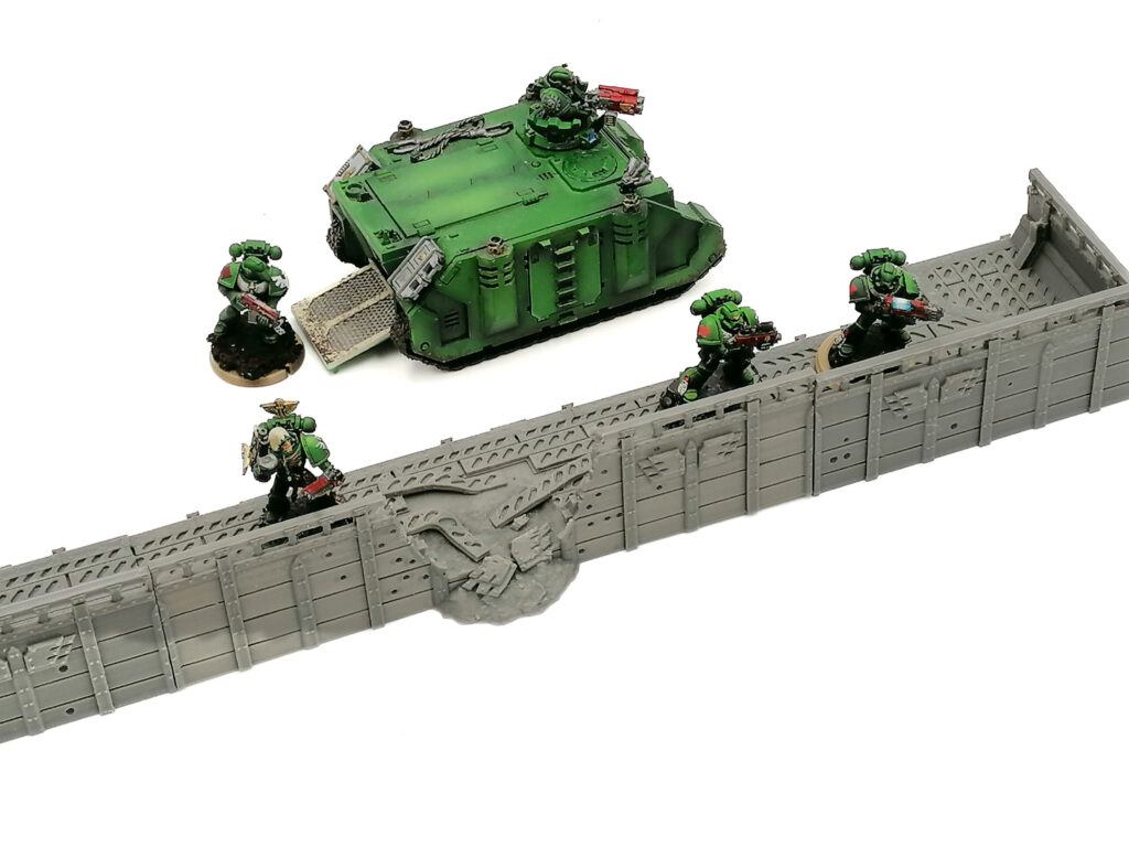 Grey 3D-printed sci-fi defensive wall with a breached section, shown with painted miniatures and a vehicle for scale