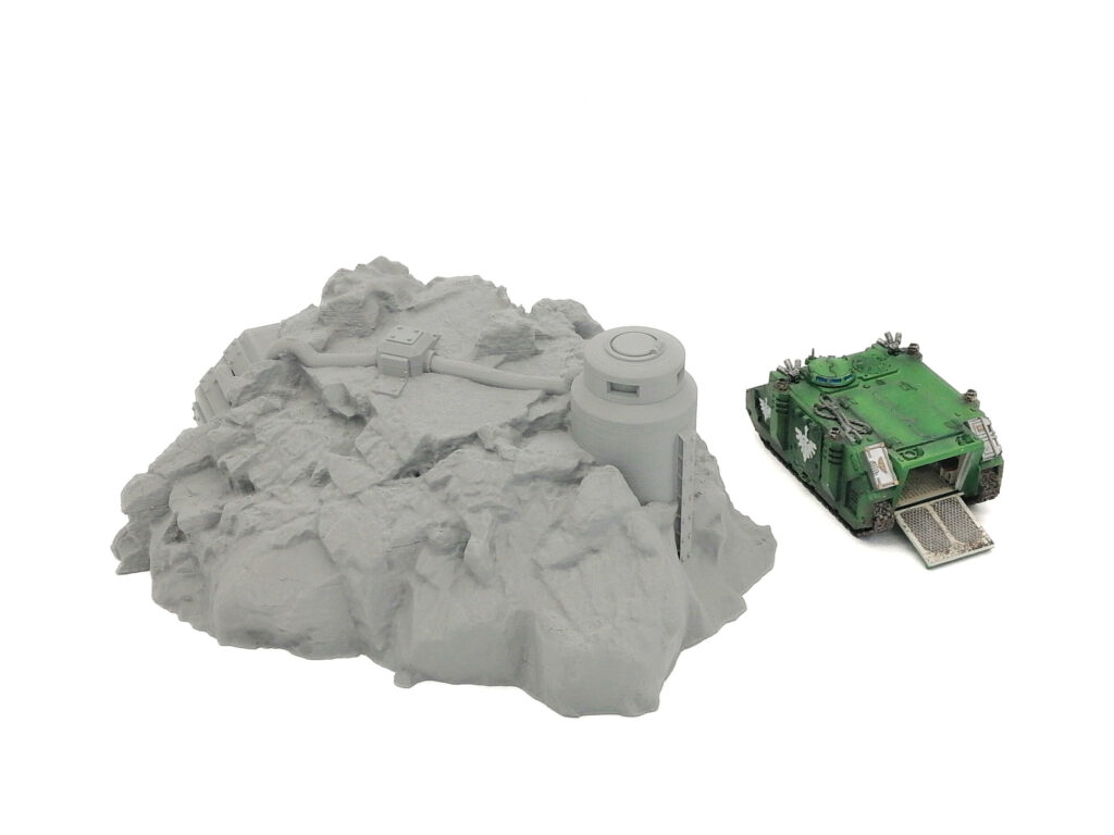 Grey unpainted 3D-printed rocky terrain with a pillbox bunker, next to a miniature green tank for scale