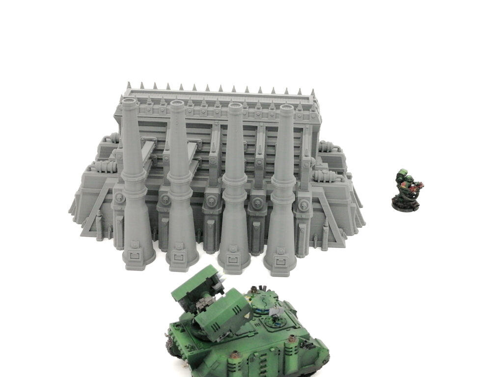 Grey 3D-printed miniature industrial building with four smokestacks, shown with a miniature tank for scale