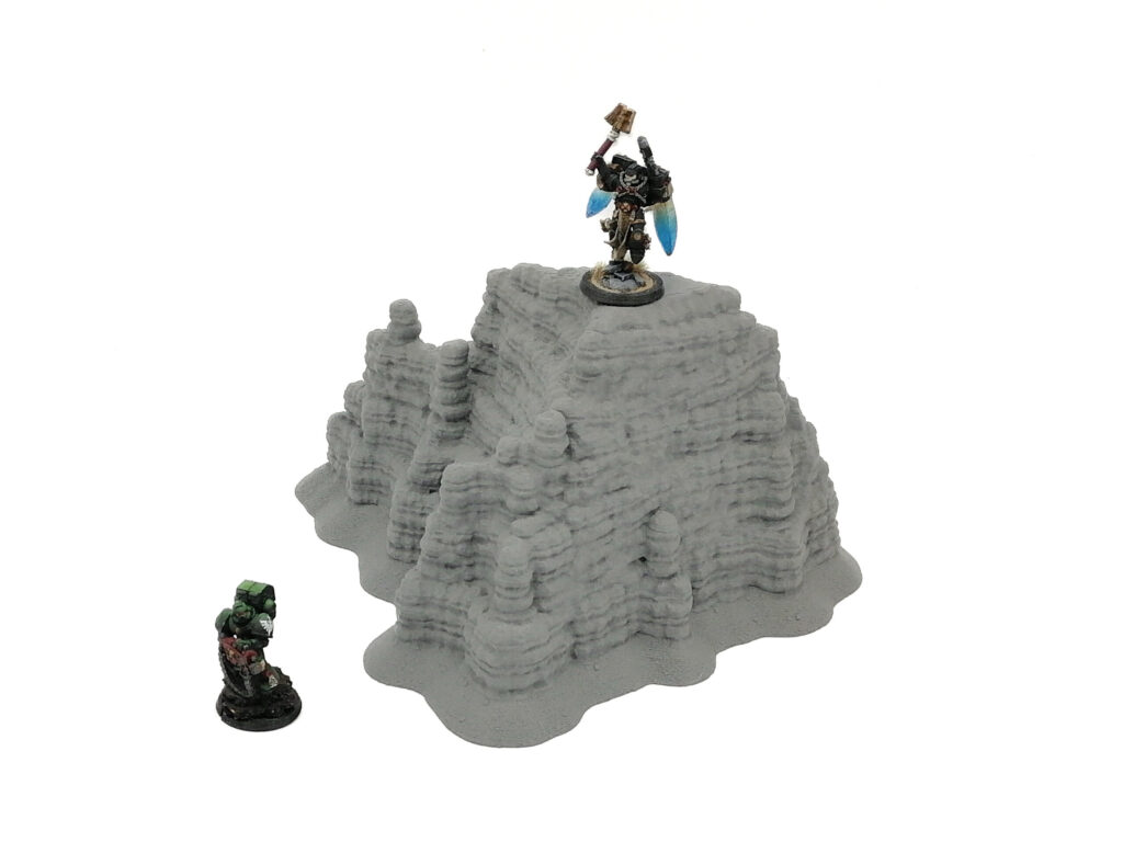 3D-printed grey rocky hill terrain for tabletop wargaming, shown with miniature figures to demonstrate scale