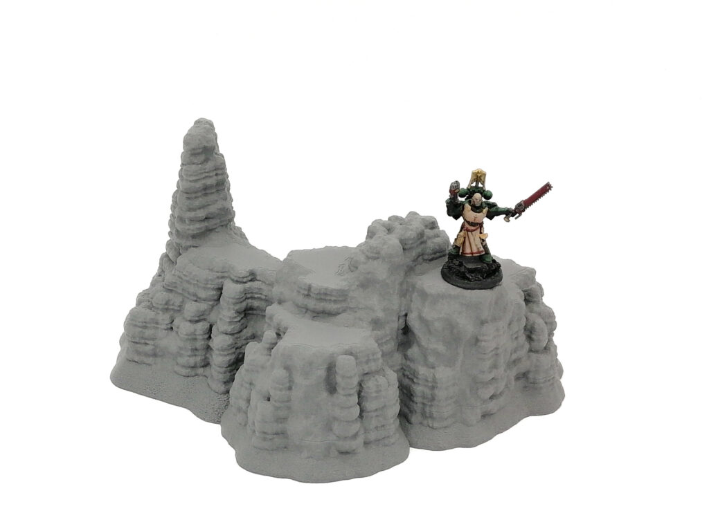 3D-printed grey rocky cliff terrain piece for tabletop gaming, shown with a painted miniature figure for scale