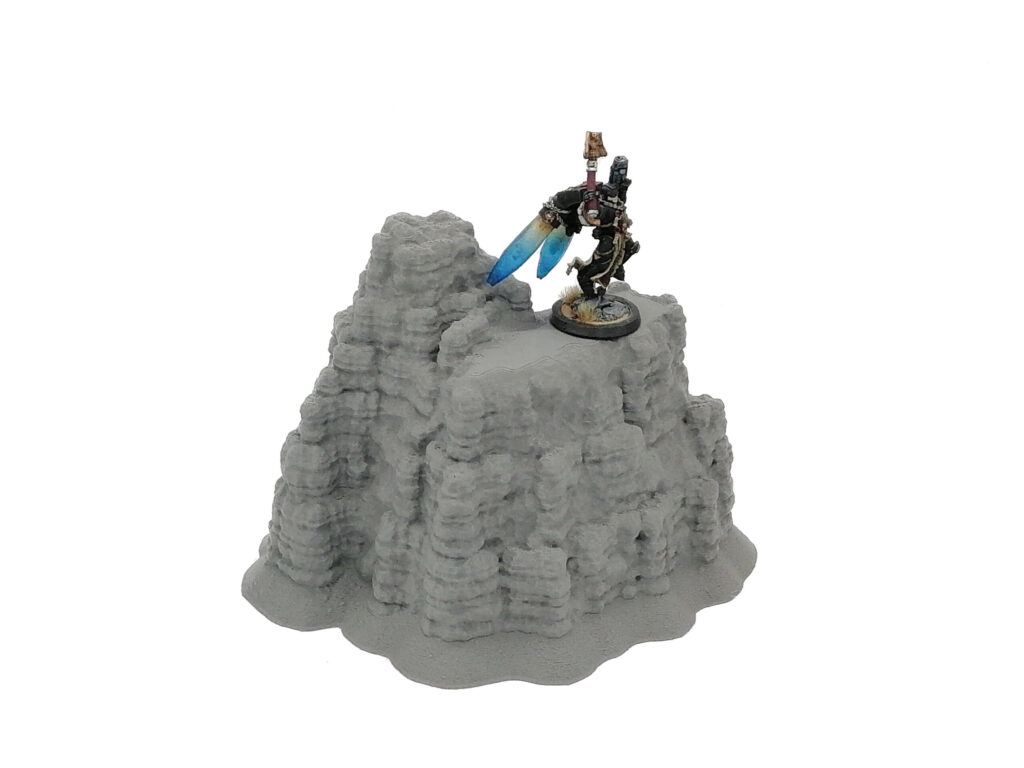 3D-printed grey rocky outcrop tabletop gaming terrain, shown with a miniature figure on top for scale