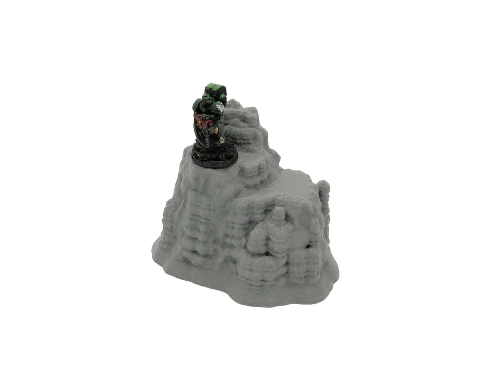 Grey 3D-printed rocky outcrop terrain miniature for tabletop gaming, shown with a painted sci-fi figure for scale