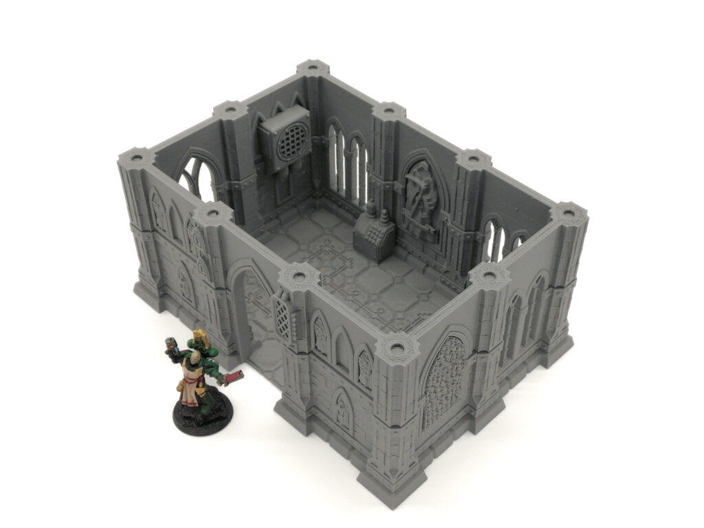 Grey 3D printed gothic building terrain for tabletop gaming, shown with a painted miniature figure for scale