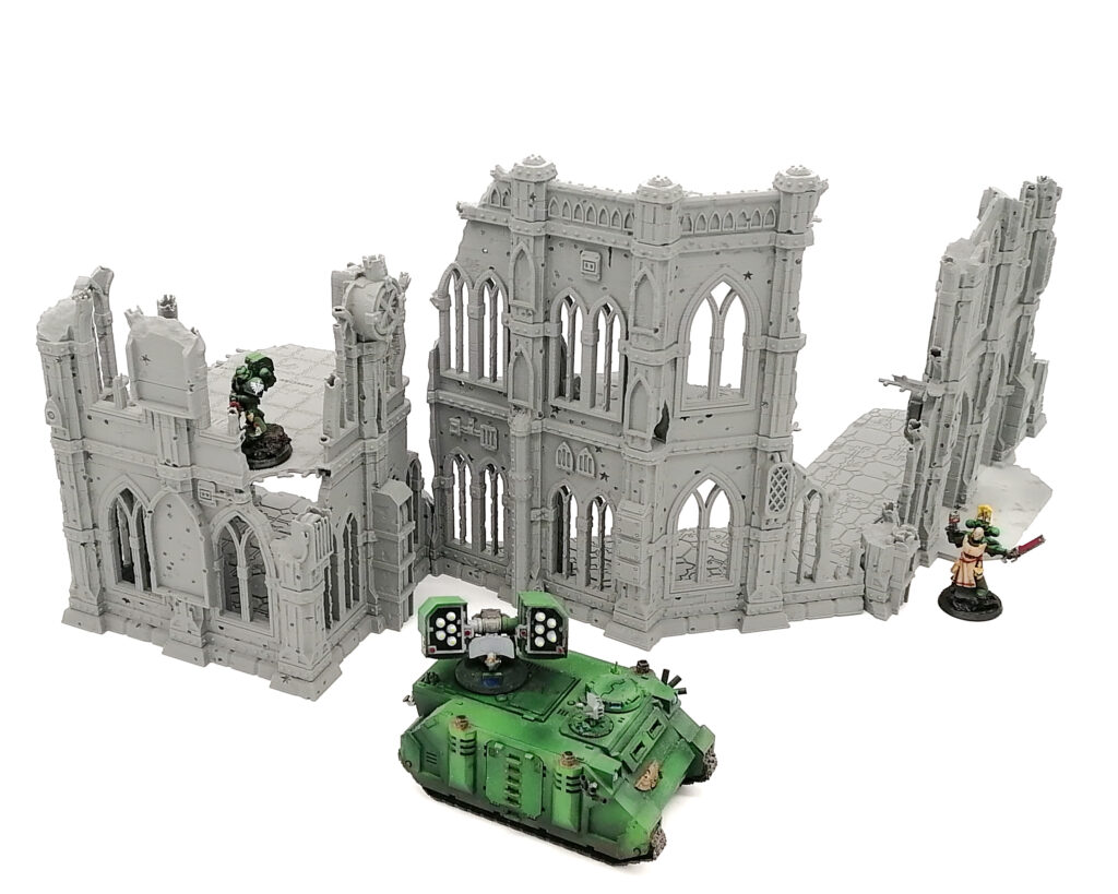 Grey 3D-printed gothic ruin terrain set for tabletop gaming, shown with a green miniature tank and soldiers