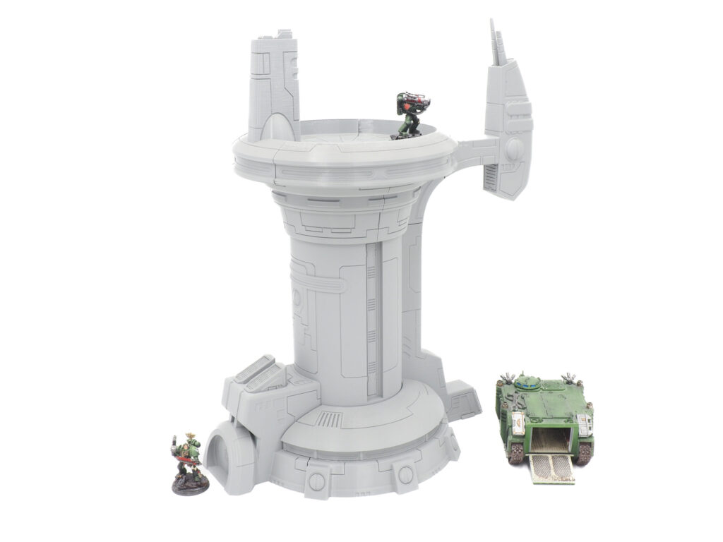 Grey 3D-printed sci-fi tower terrain for tabletop gaming, shown with miniatures and a tank for scale