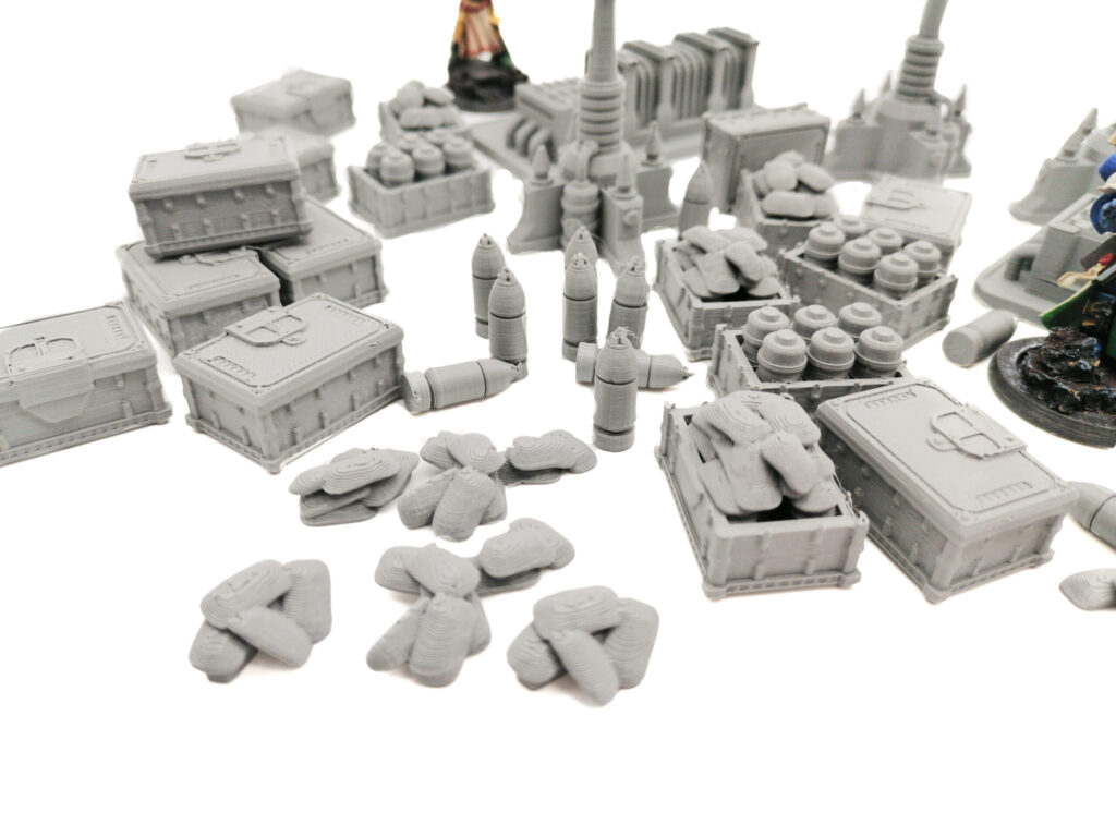 Grey 3D-printed tabletop wargaming terrain miniatures including crates, sandbags, and artillery shells