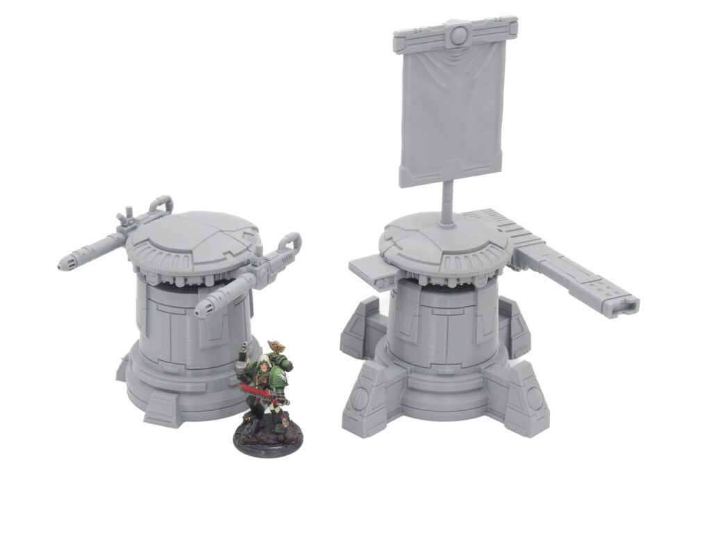 Two grey 3D-printed sci-fi turrets, one with cannons and one with a banner, next to a painted miniature for scale