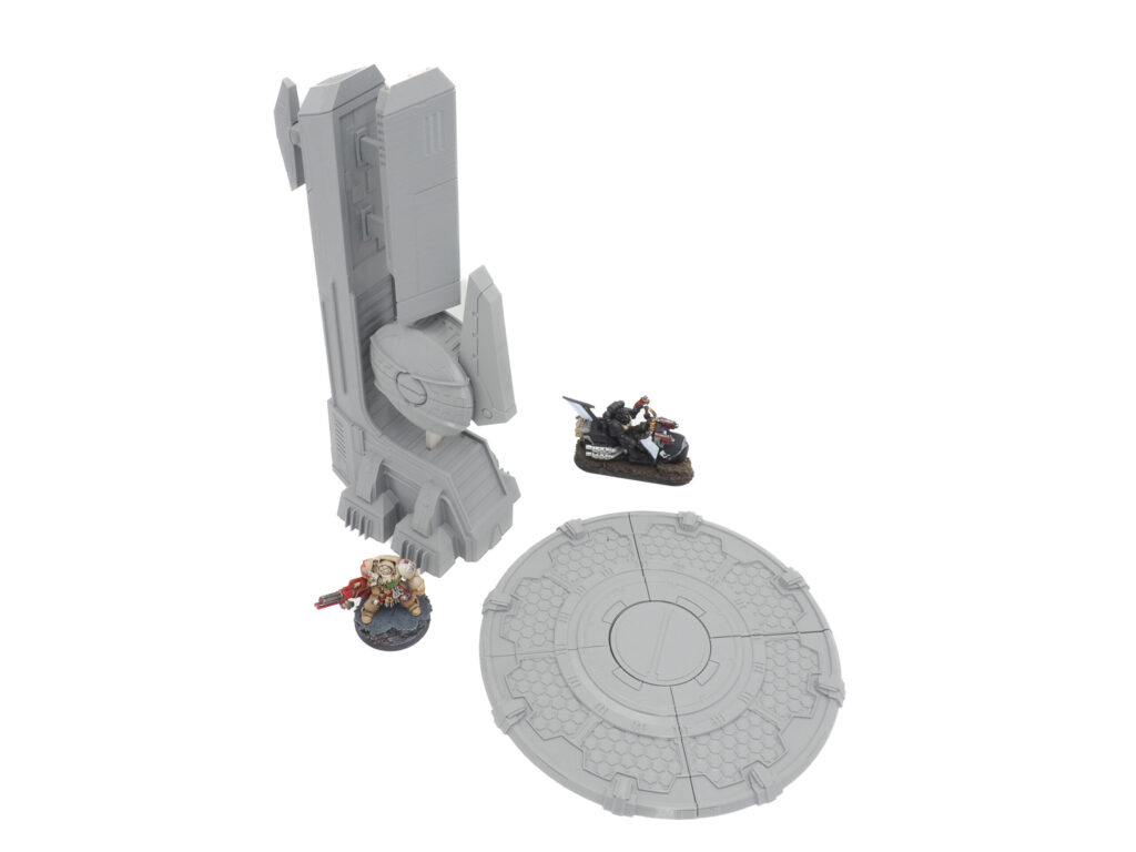 Grey 3D-printed sci-fi wargaming terrain, featuring a tall tower and circular pad shown with miniatures for scale