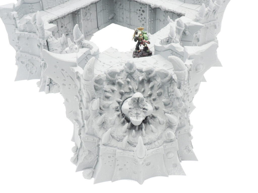 Unpainted grey 3D printed sci-fi fortress terrain with a toothy maw design and a small miniature on top for scale