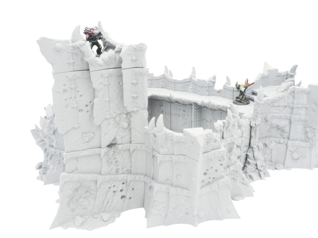 Grey 3D-printed tabletop wargaming fortress wall terrain with spikes, shown with miniature figures for scale