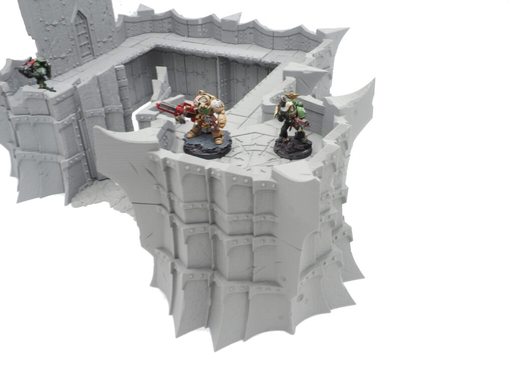 Grey 3D-printed modular fortress wall terrain for tabletop wargames, shown with painted miniatures for scale