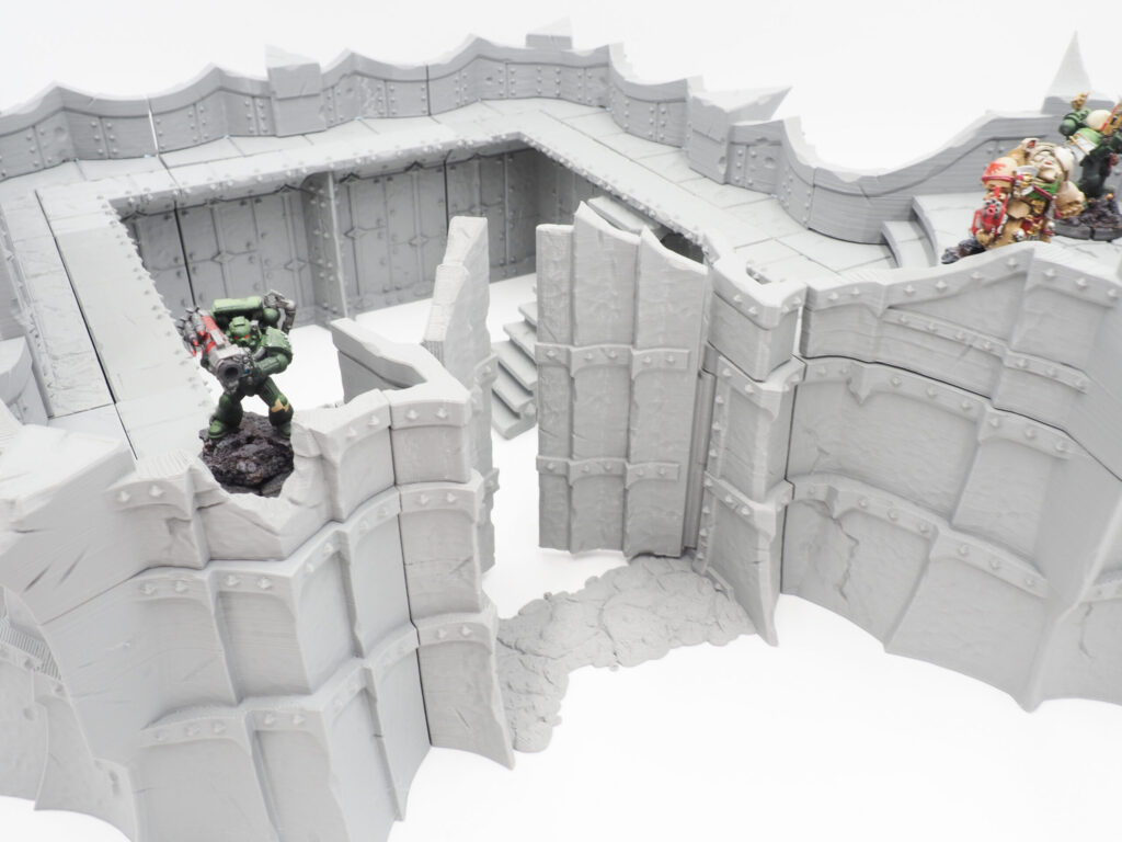 Grey 3D-printed modular fortress walls and gate for tabletop gaming, shown with painted miniatures