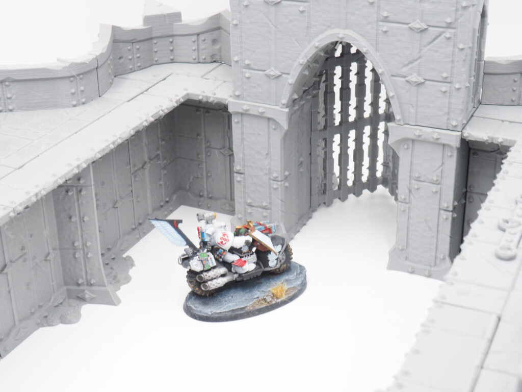 Grey 3D-printed modular fortress walls and gatehouse, shown with a painted miniature motorcycle rider for scale