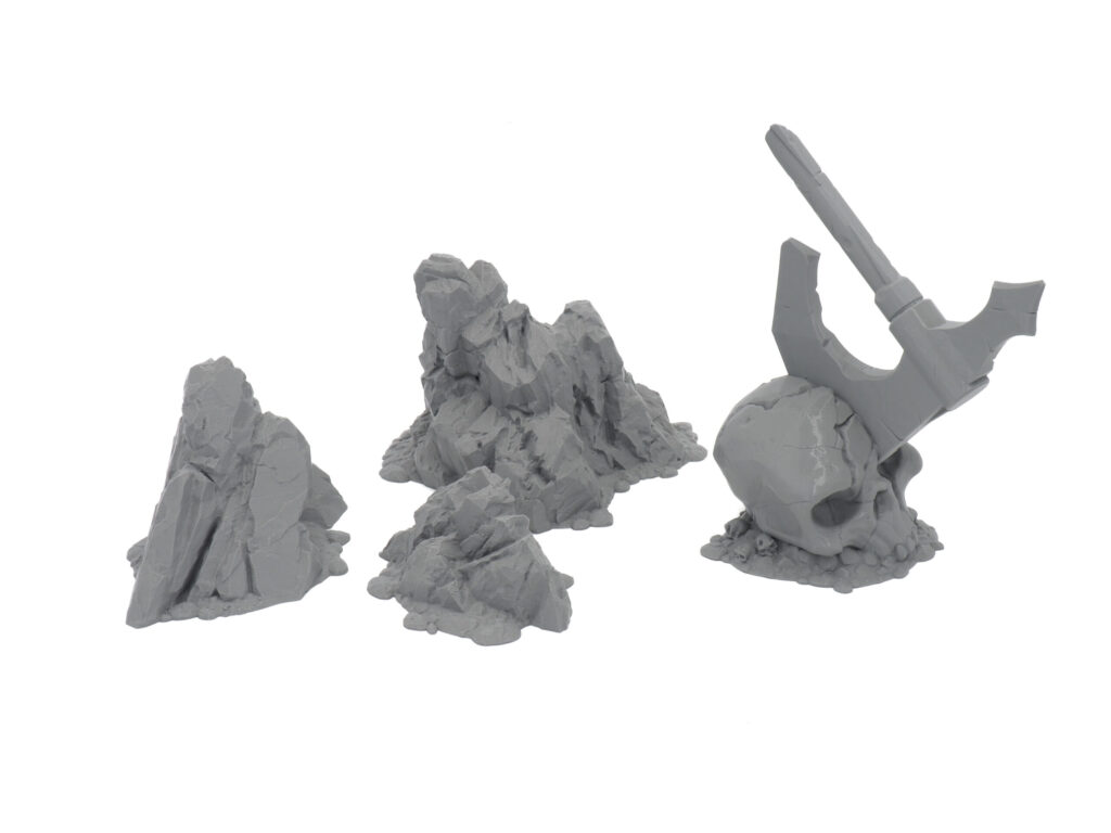 Grey 3D-printed tabletop gaming terrain pieces featuring three rocky formations and a skull with an embedded battleaxe
