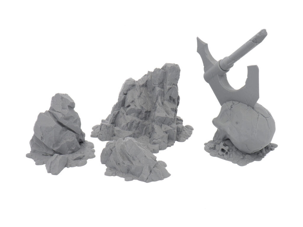 Grey 3D-printed tabletop terrain miniatures featuring rocky outcrops and a skull with an axe embedded in it