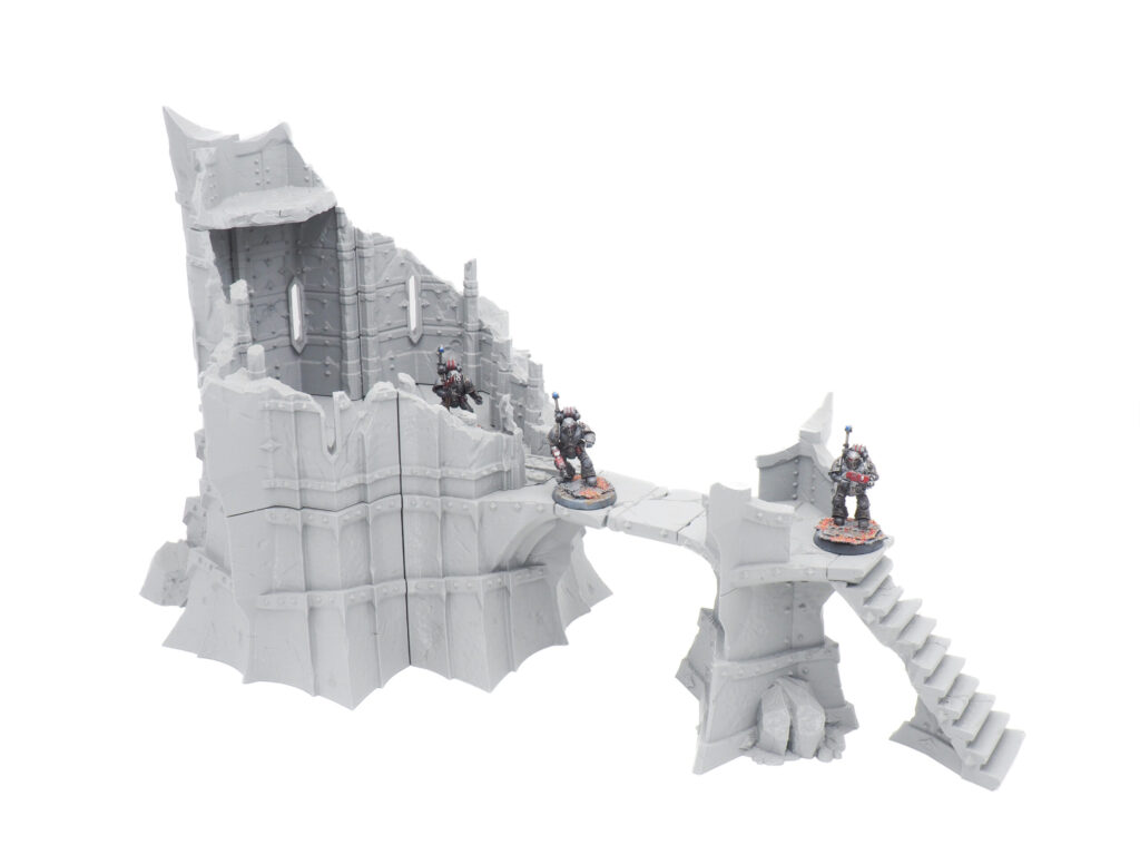 Grey 3D-printed gothic ruined tower terrain with a connecting bridge and stairs, shown with miniatures for scale