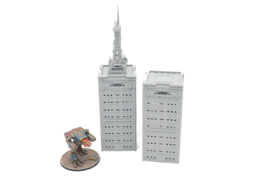 Two grey 3D-printed miniature skyscraper buildings, one with an antenna tower, shown next to a painted mech for scale