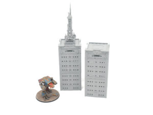 Two grey 3D-printed miniature skyscraper buildings, one with an antenna tower, shown next to a painted mech for scale