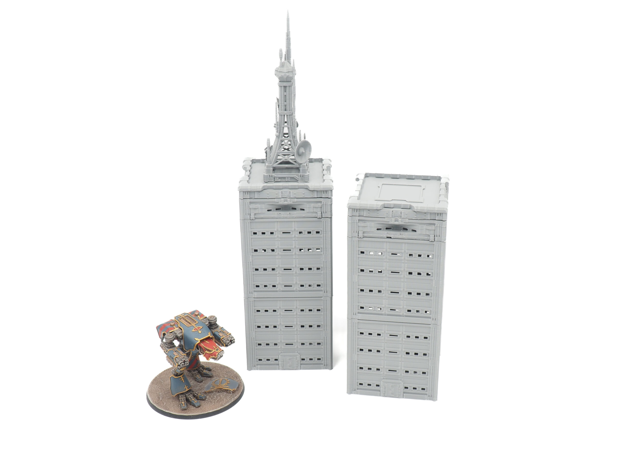 Two grey 3D-printed miniature skyscraper buildings, one with an antenna tower, shown next to a painted mech for scale