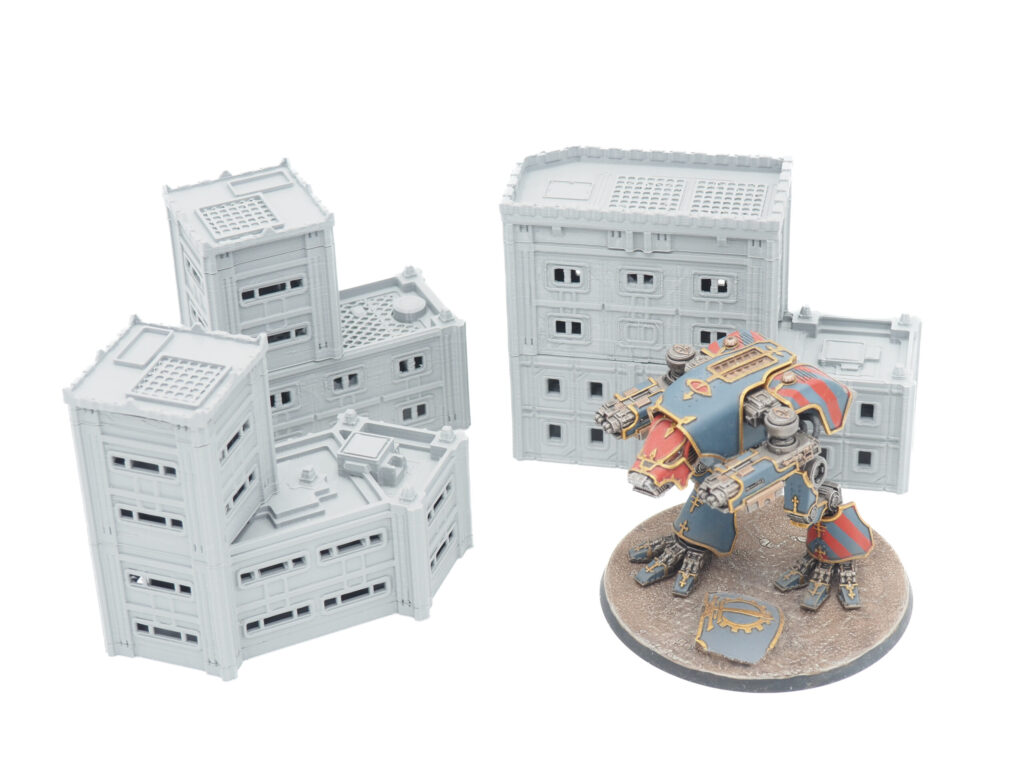 Unpainted grey 3D-printed miniature buildings for tabletop gaming, shown next to a painted mech figure for scale