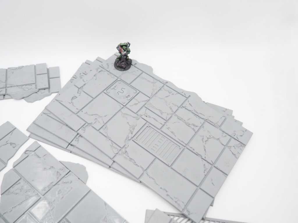 Grey 3D printed modular floor tiles with cracked details, shown with a painted miniature gaming figure for scale