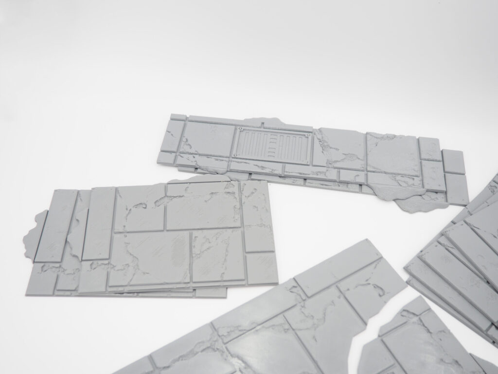 Grey 3D-printed tabletop terrain pieces depicting cracked stone pavement, featuring a rectangular drain grate