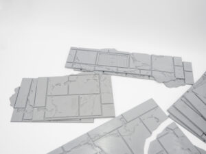 Grey 3D-printed tabletop terrain pieces depicting cracked stone pavement, featuring a rectangular drain grate