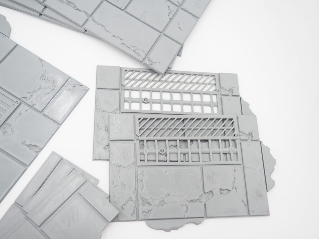 Grey 3D-printed plastic tabletop terrain tiles featuring textured pavements and drainage grates