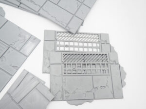 Grey 3D-printed plastic tabletop terrain tiles featuring textured pavements and drainage grates