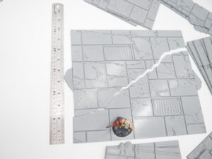 Grey 3D-printed modular stone tiles for tabletop gaming, shown with a ruler and miniature figurine for scale