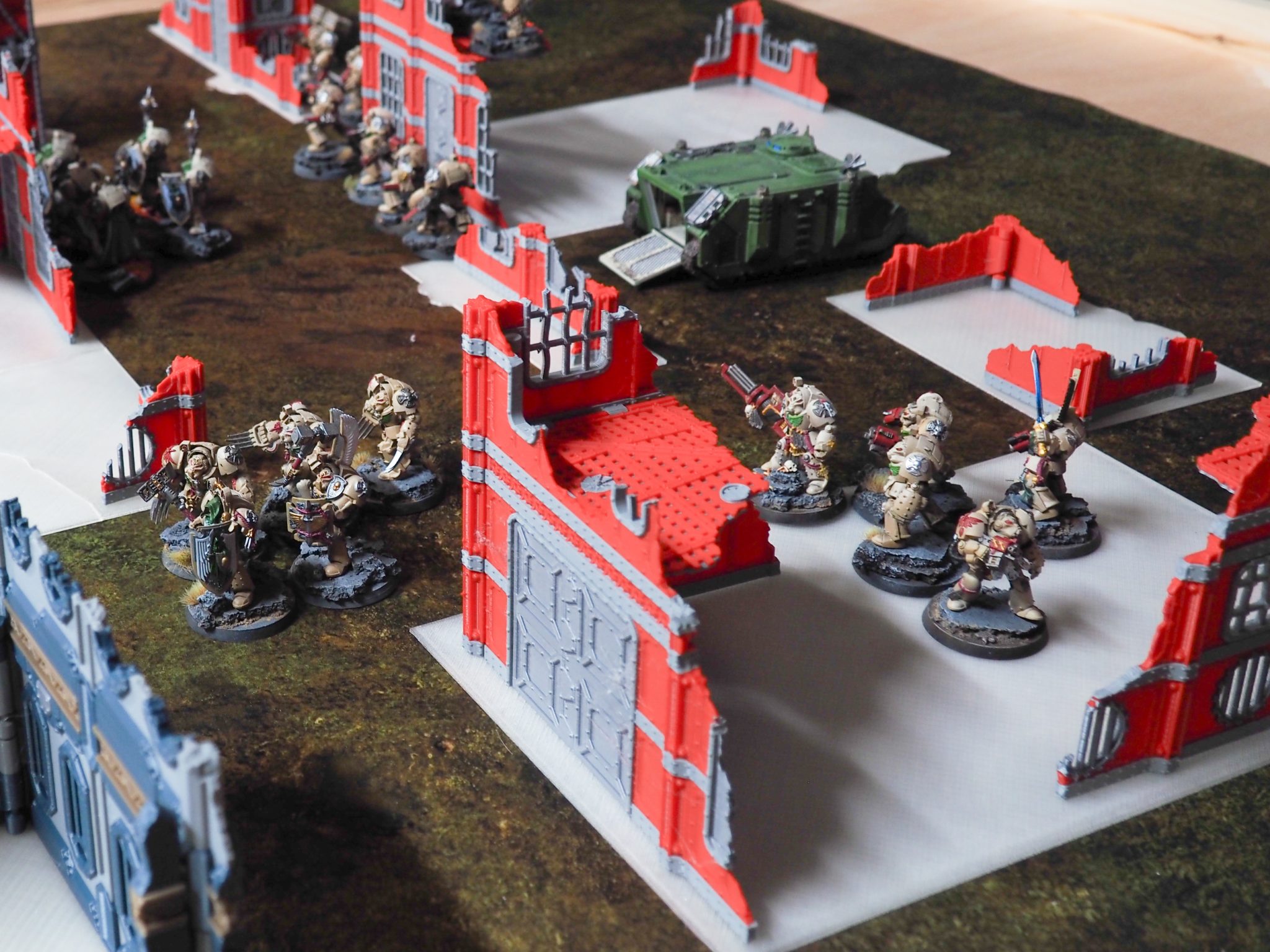 3D-printed red and grey ruined building terrain pieces set up on a tabletop gaming mat with miniatures