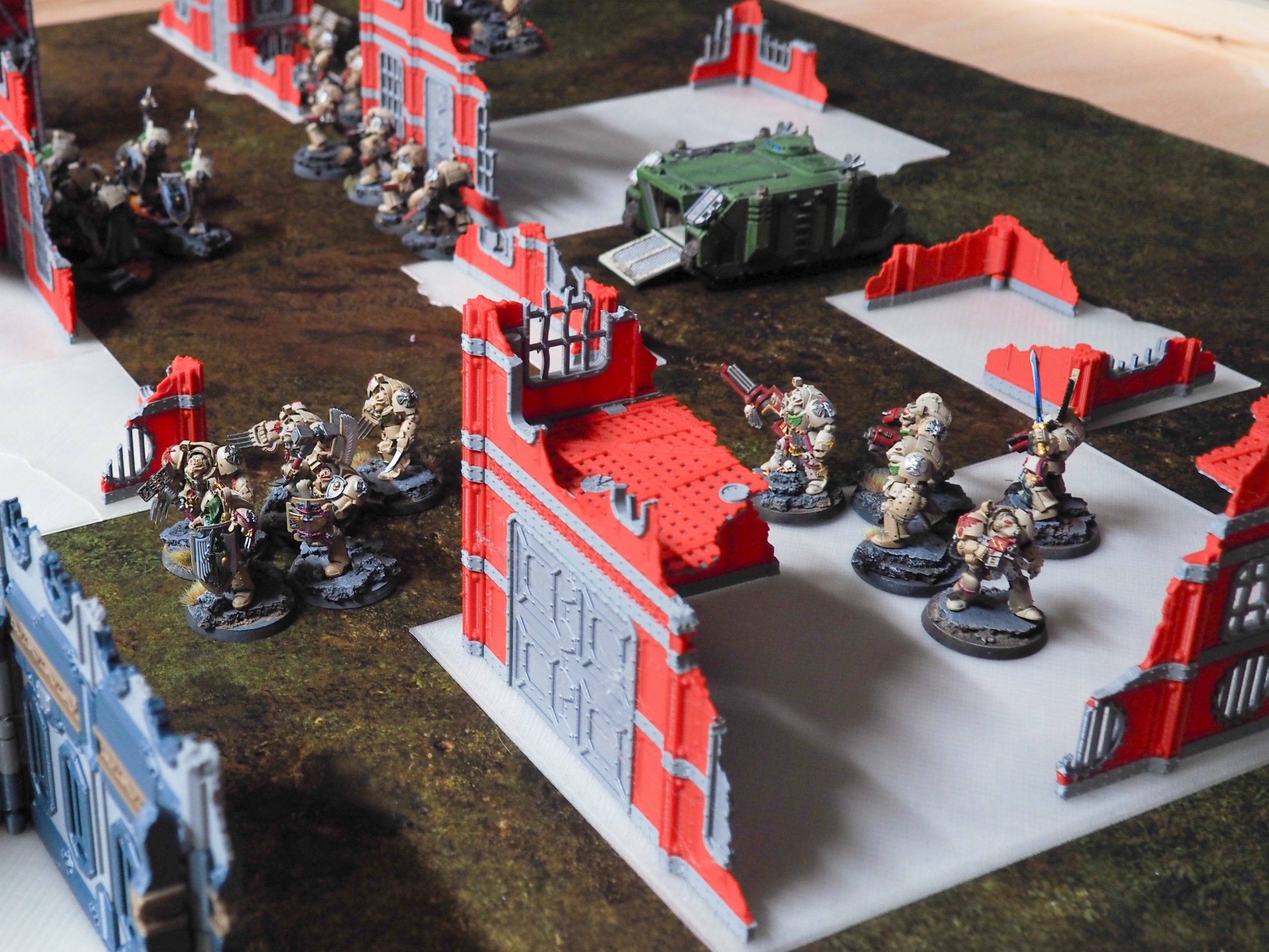 3D-printed red and grey ruined building terrain pieces set up on a tabletop gaming mat with miniatures