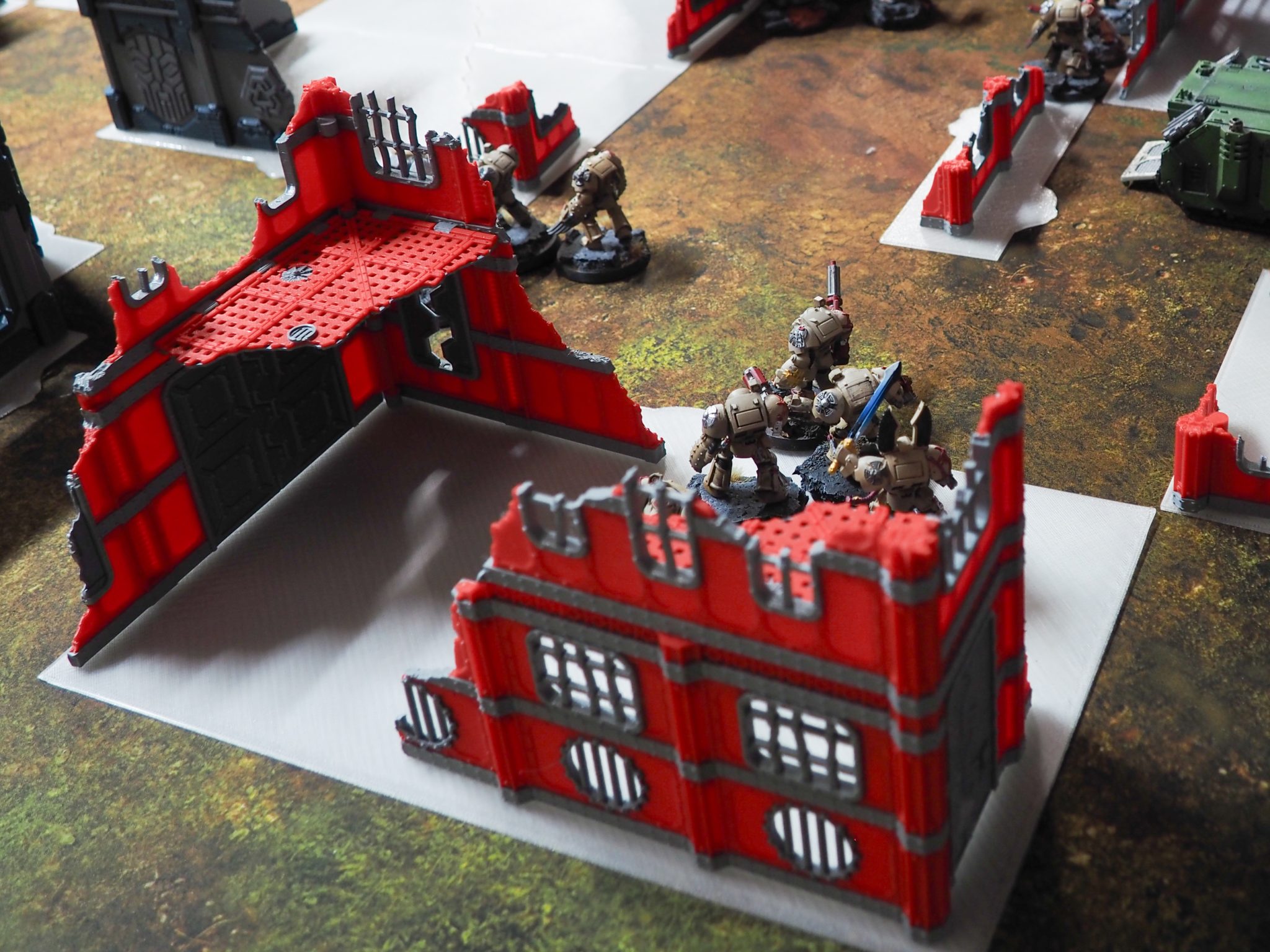 Red and grey 3D printed ruined building terrain set up with miniature figures on a tabletop wargaming mat
