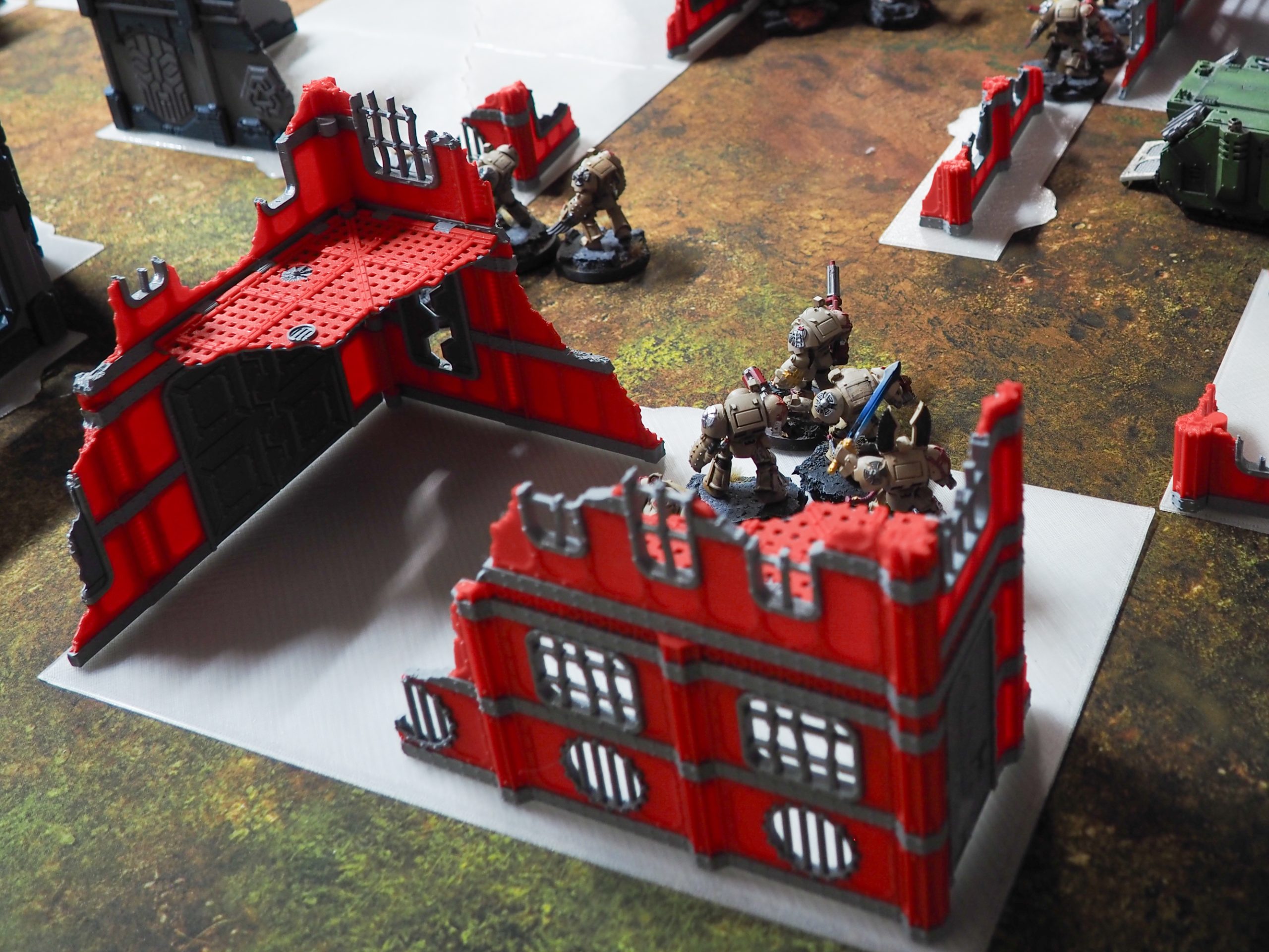 Red and grey 3D printed ruined building terrain set up with miniature figures on a tabletop wargaming mat