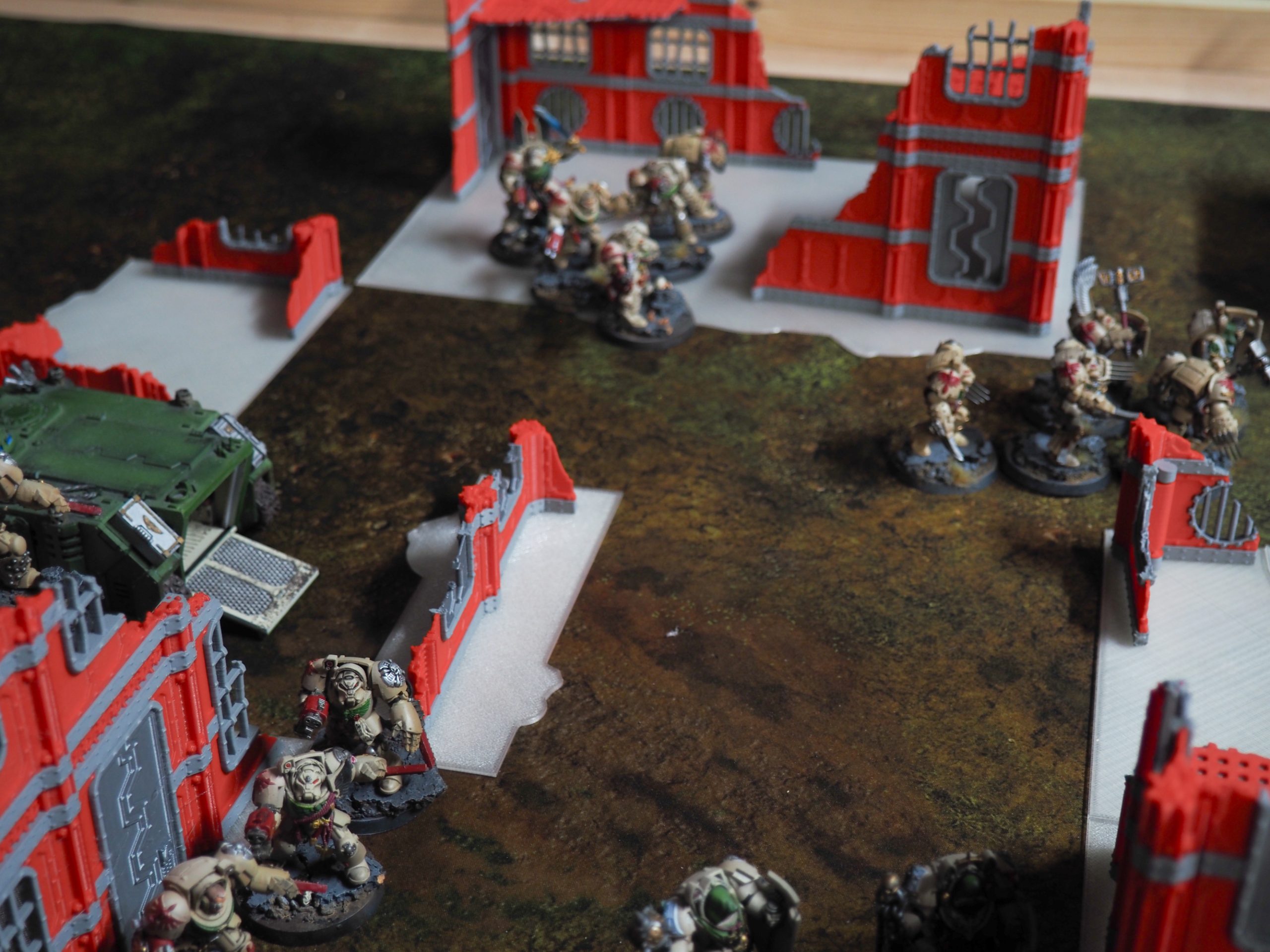 3D-printed red and grey ruined building terrain pieces set up on a tabletop gaming mat with miniatures