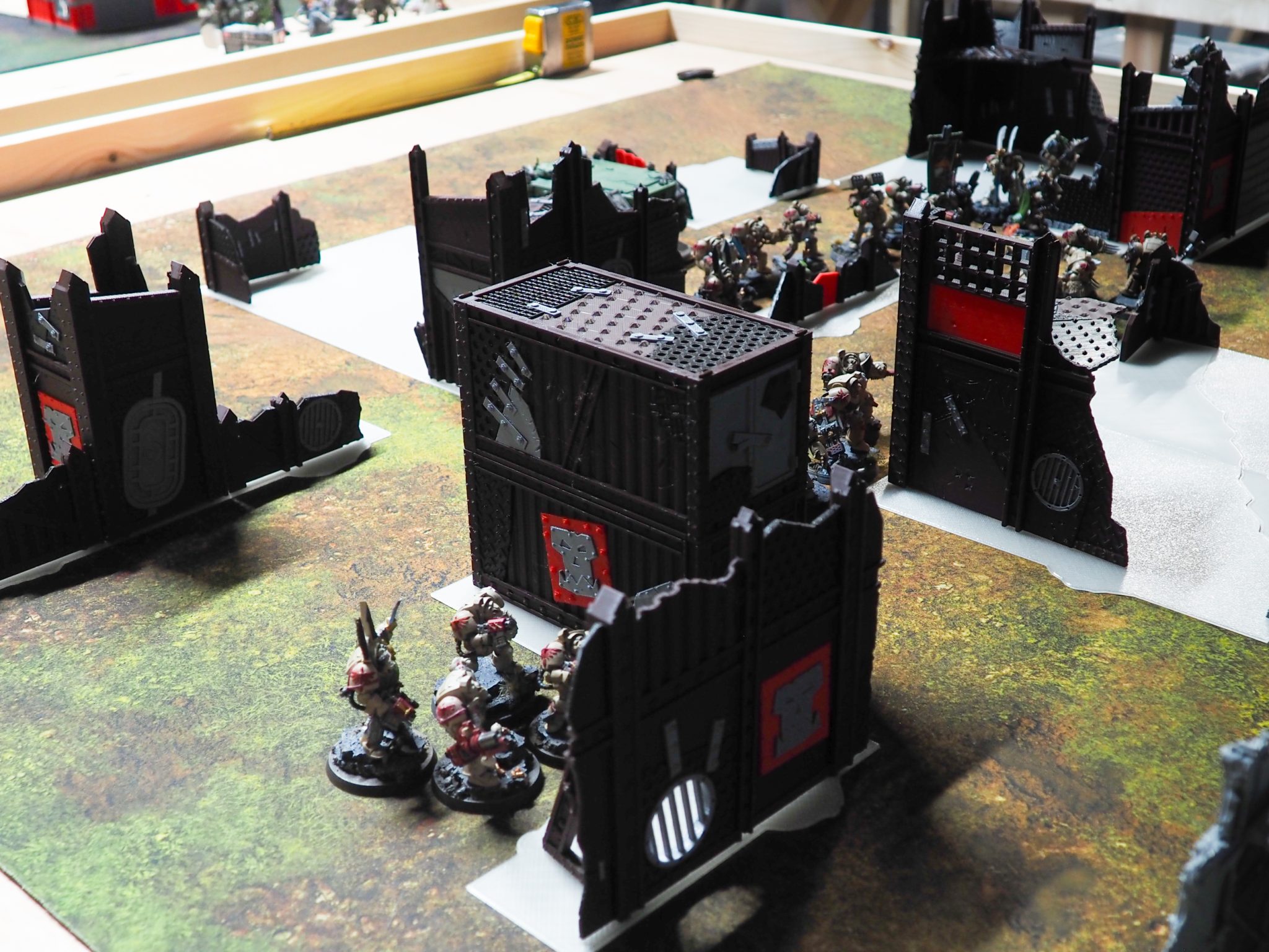 3D-printed tabletop wargaming terrain with dark industrial barricades and stacked containers set up with miniatures