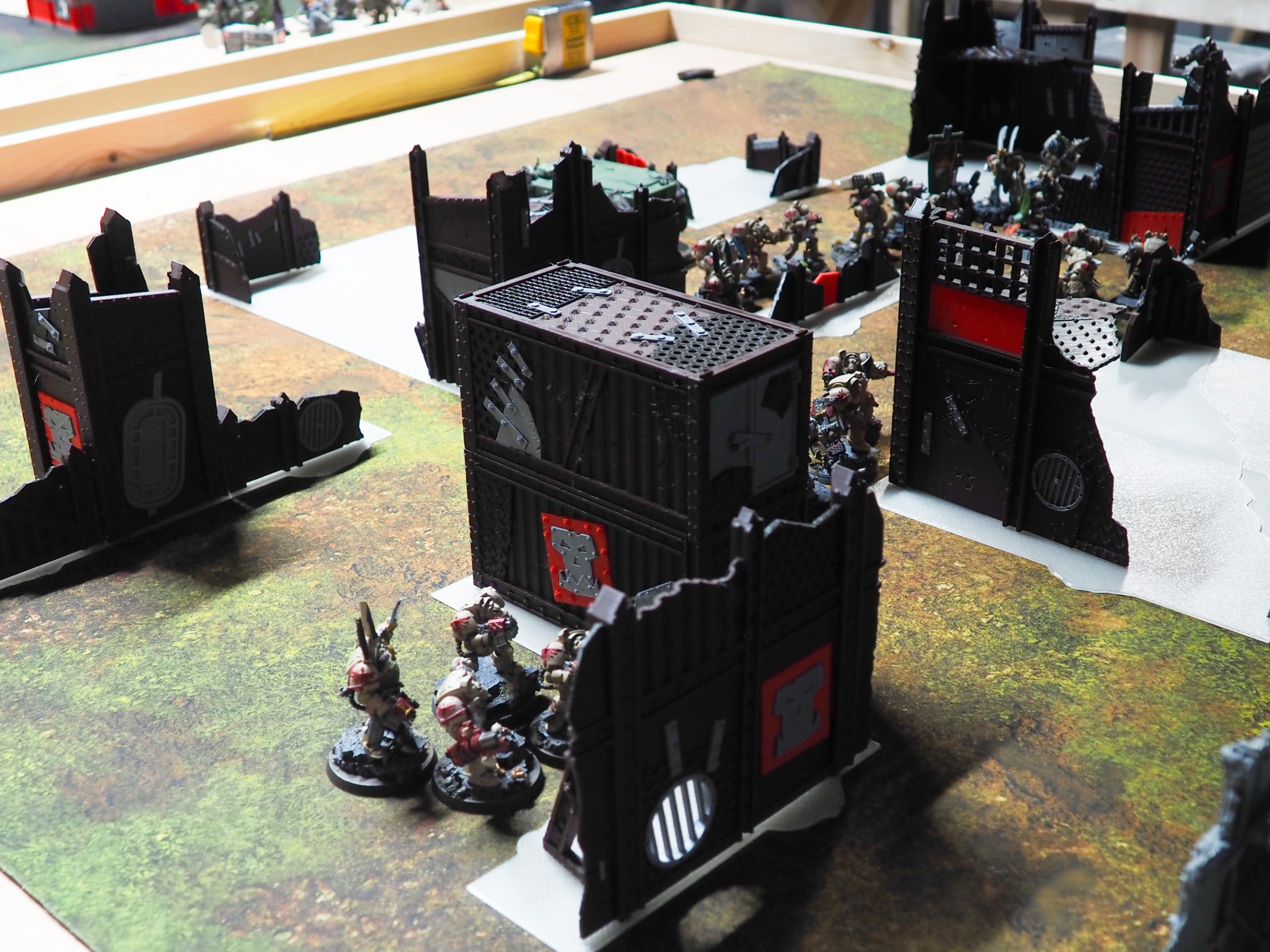 3D-printed tabletop wargaming terrain with dark industrial barricades and stacked containers set up with miniatures