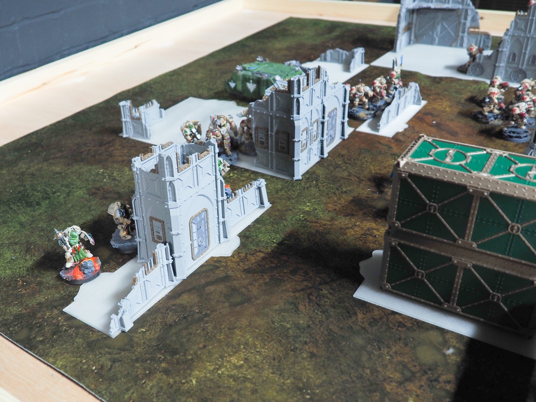 3D printed grey gothic building terrain and green containers set up on a tabletop wargaming mat