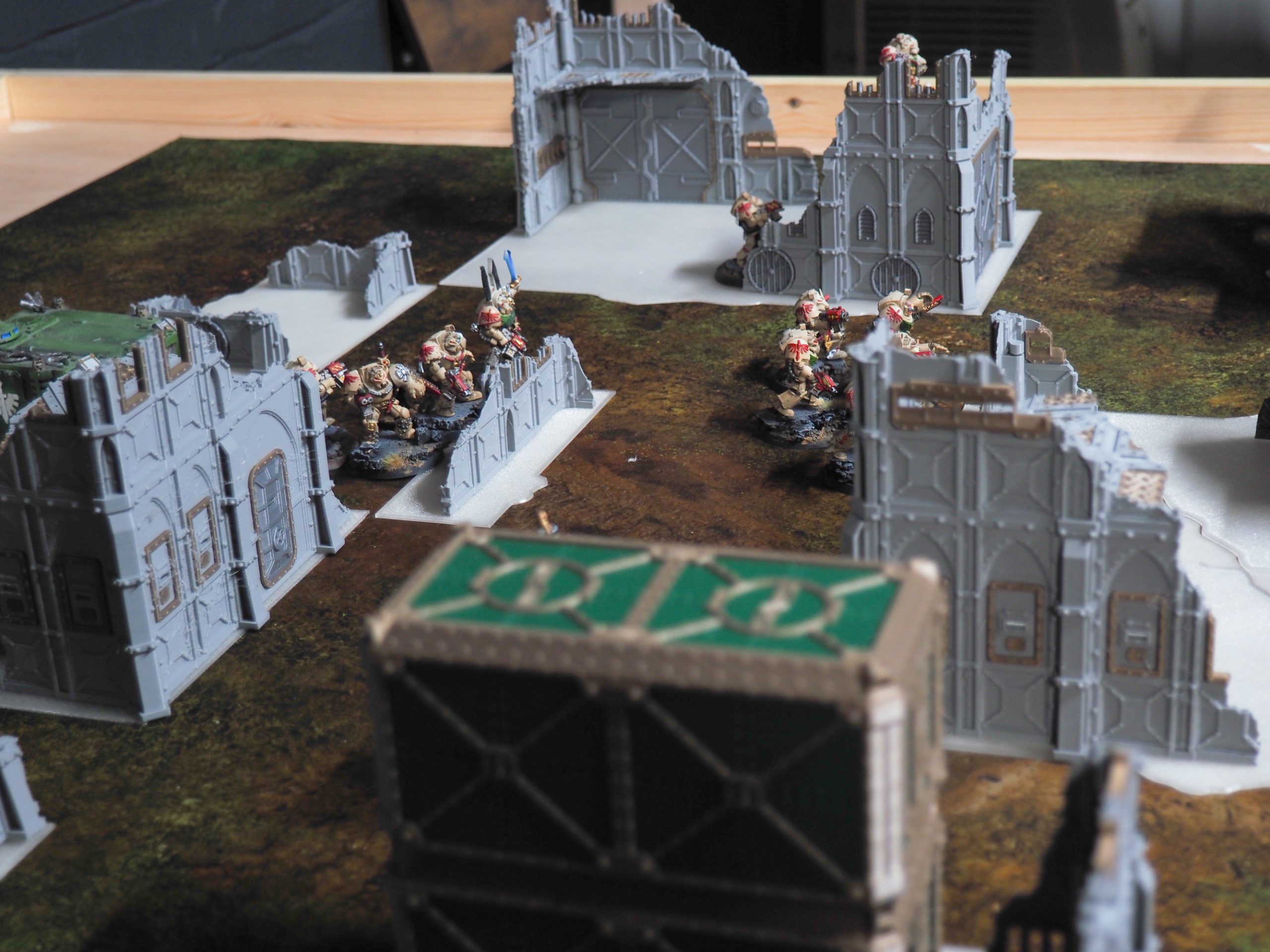 3D-printed grey gothic ruined buildings and barricades set up on a tabletop wargaming mat with miniature figures