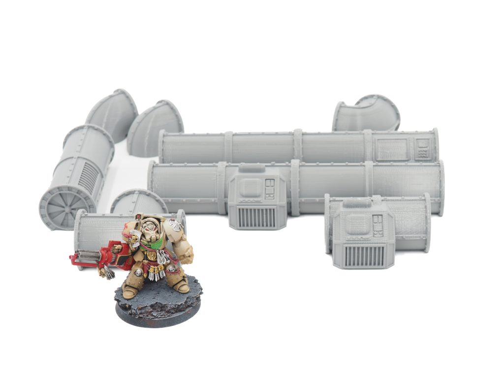 Grey 3D-printed modular pipe terrain pieces shown with a painted miniature for scale